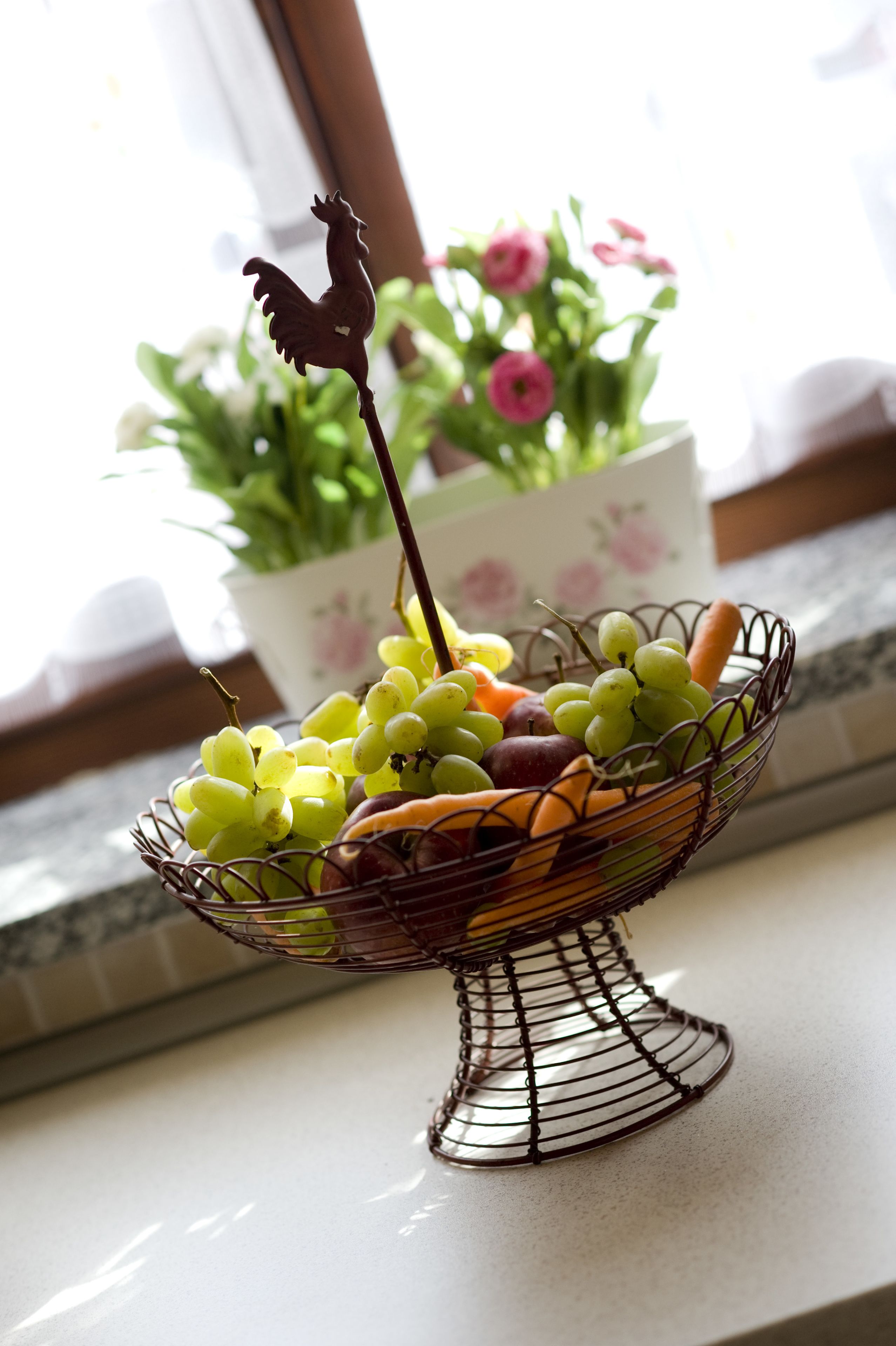 A fruit basket on a kitchen counter with a rooster decoration, a potted plant, and a window in the background.