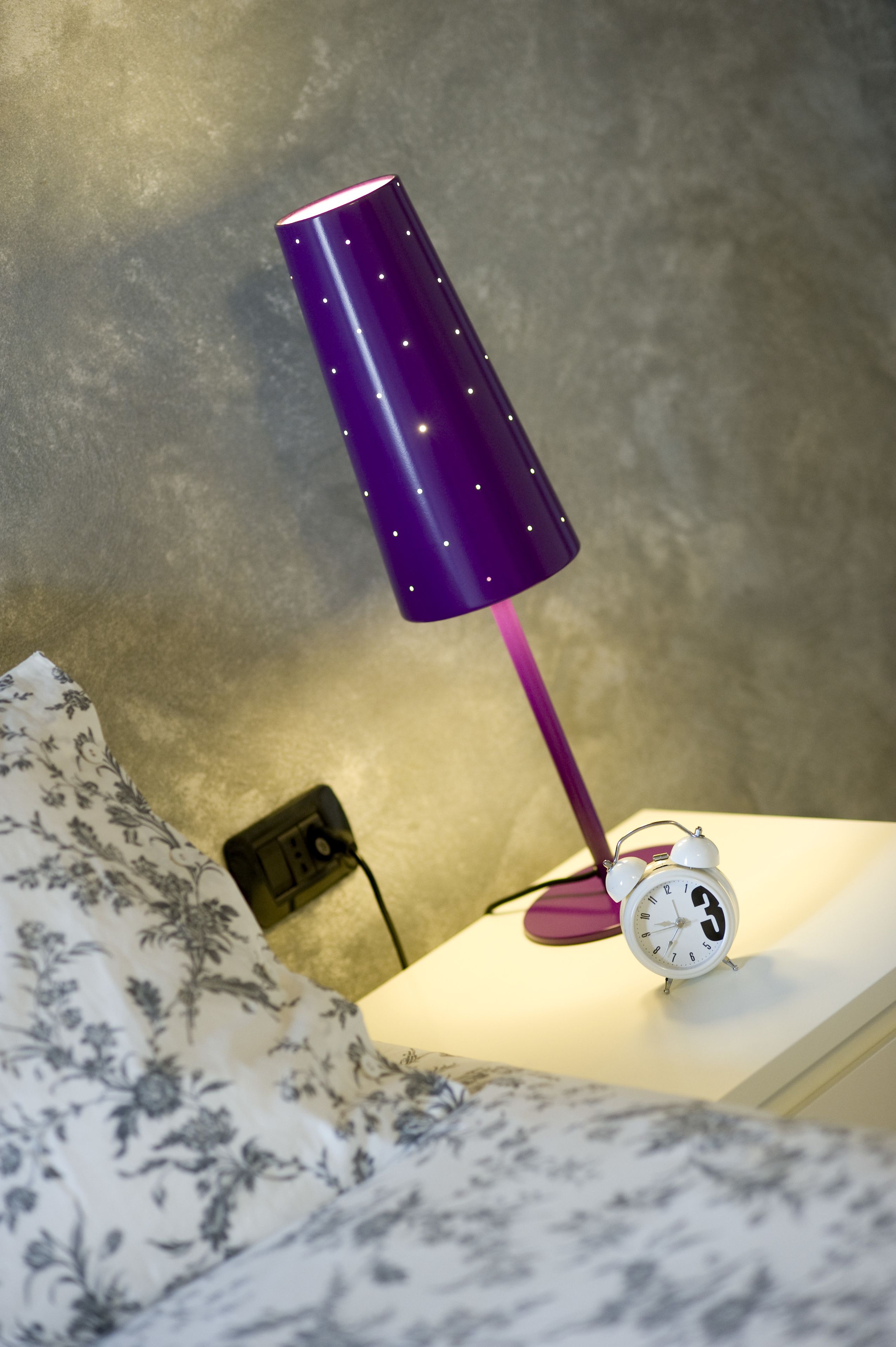 En seng med et blomstret tæppe, en lilla lampe og en hvid alarmklokke på et natbord.
