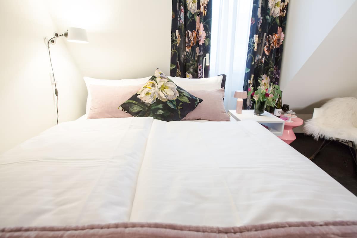 A neatly made bed with white and pink bedding, a floral pillow, a bedside table with a pink lamp, and a vase with flowers.