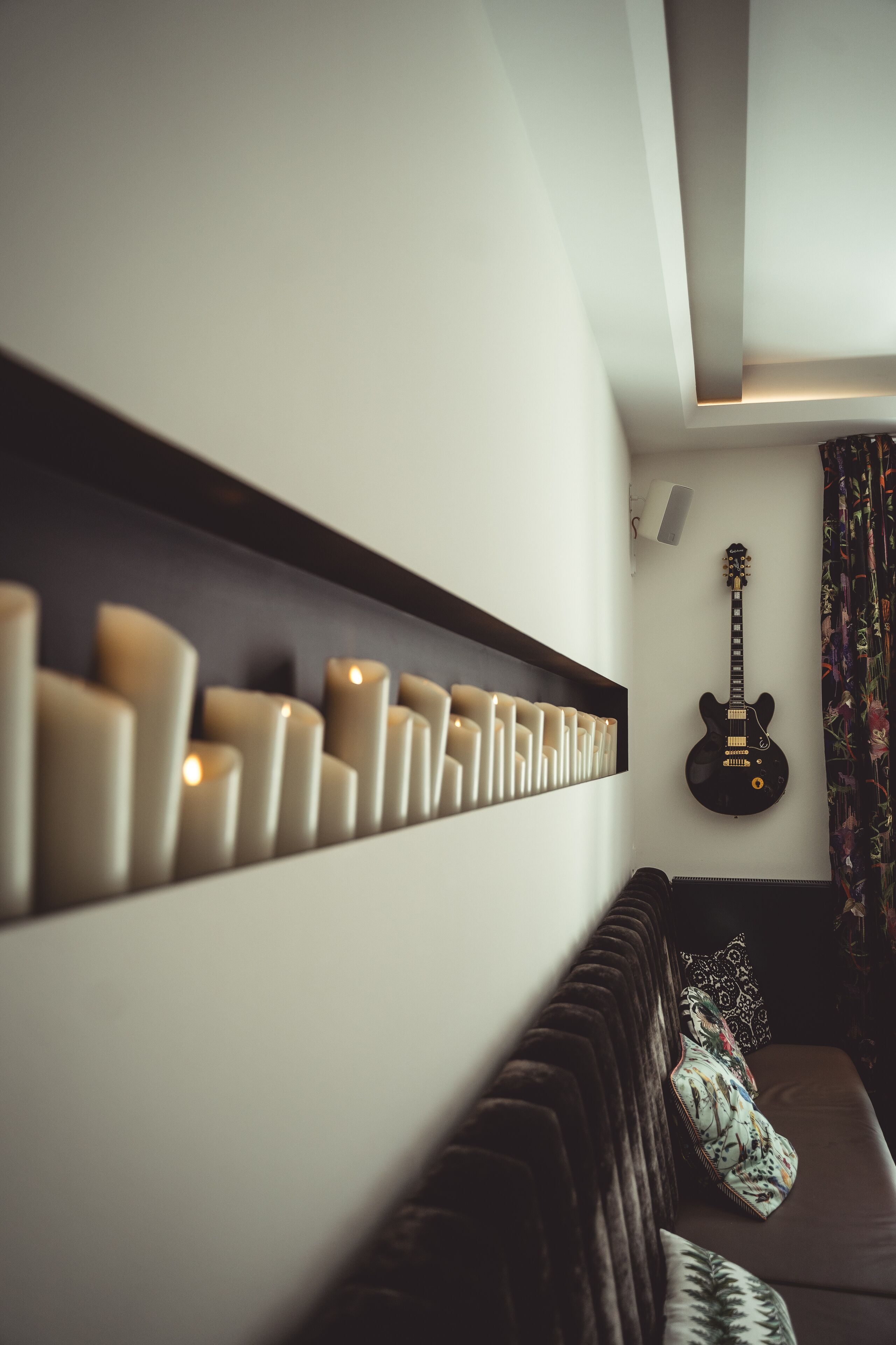 Ein Zimmer mit einer Wandablage, auf der Kerzen, eine Gitarre und ein dunkelbraunes Sofa mit gemusterten Kissen stehen.
