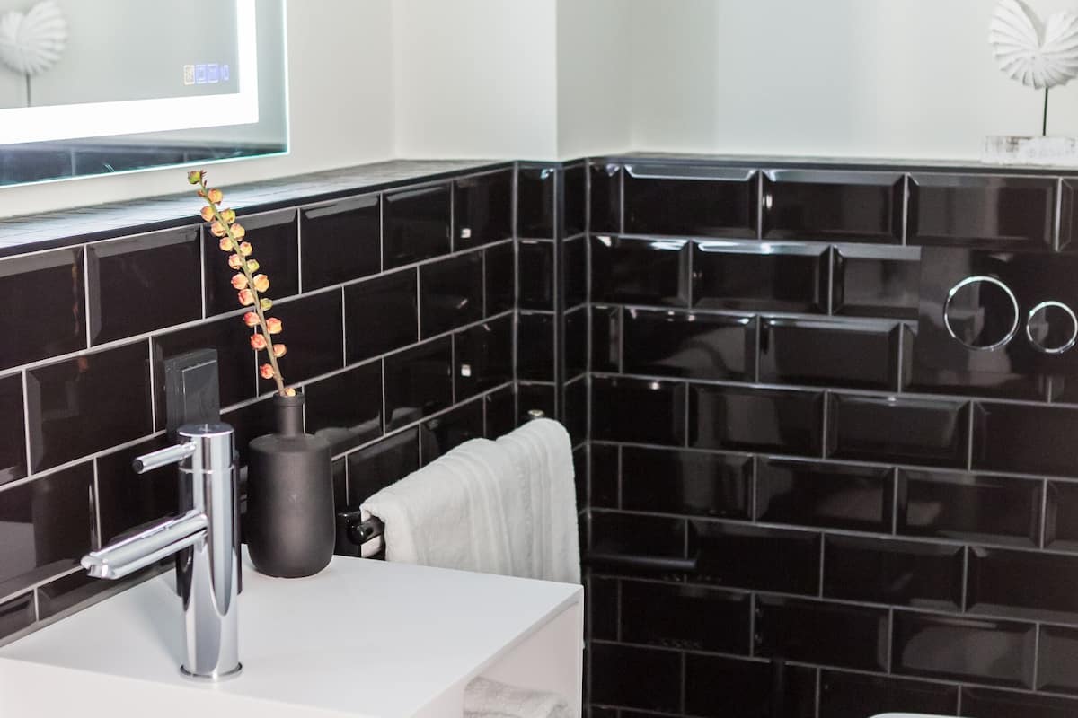 A modern bathroom with a white sink, black tiled walls, a white toilet, and a mirror with a round frame.