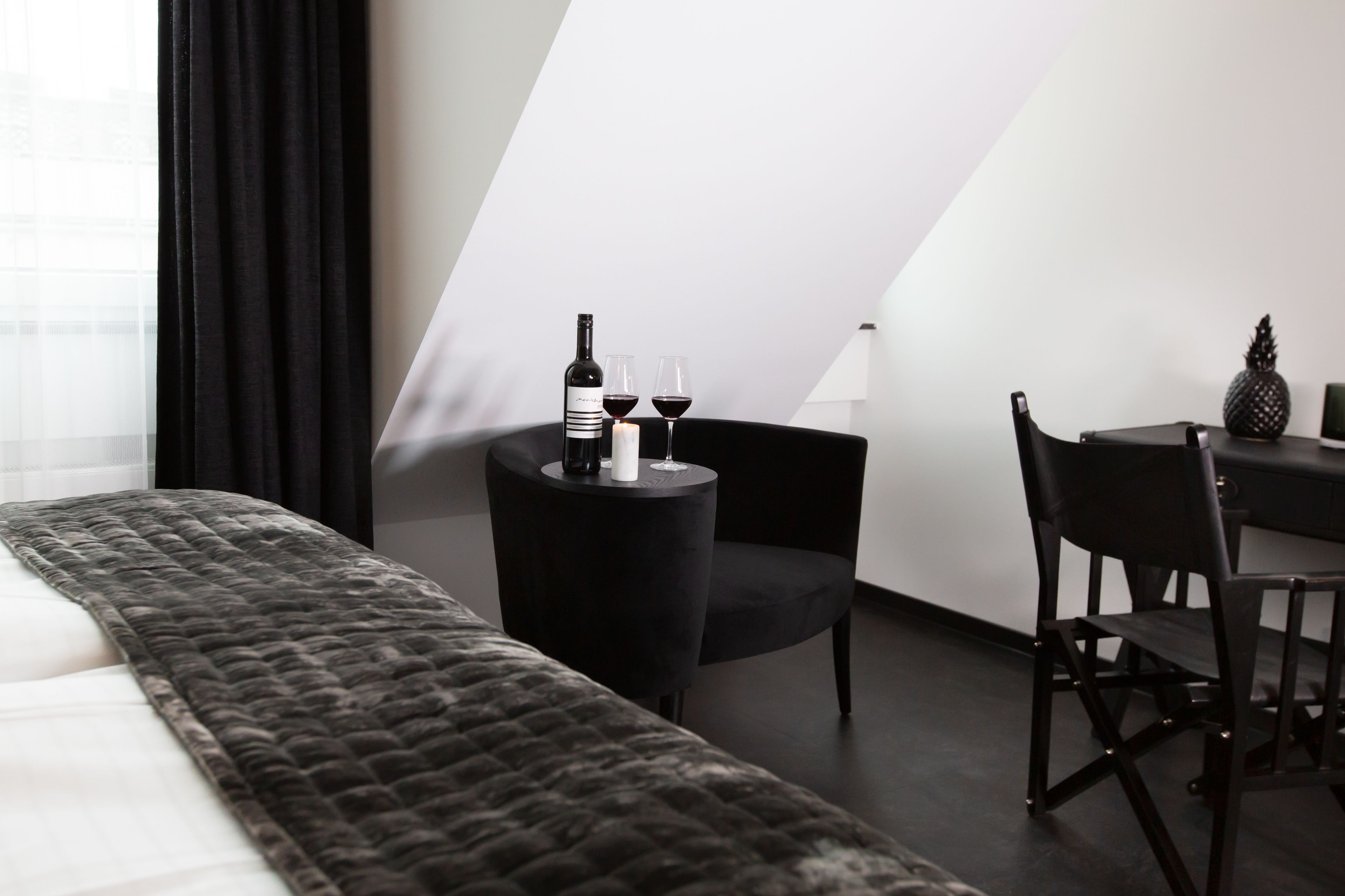 A modern hotel room with a bed, a small round table with a bottle of wine and two glasses, a chair, and a pineapple on the table.