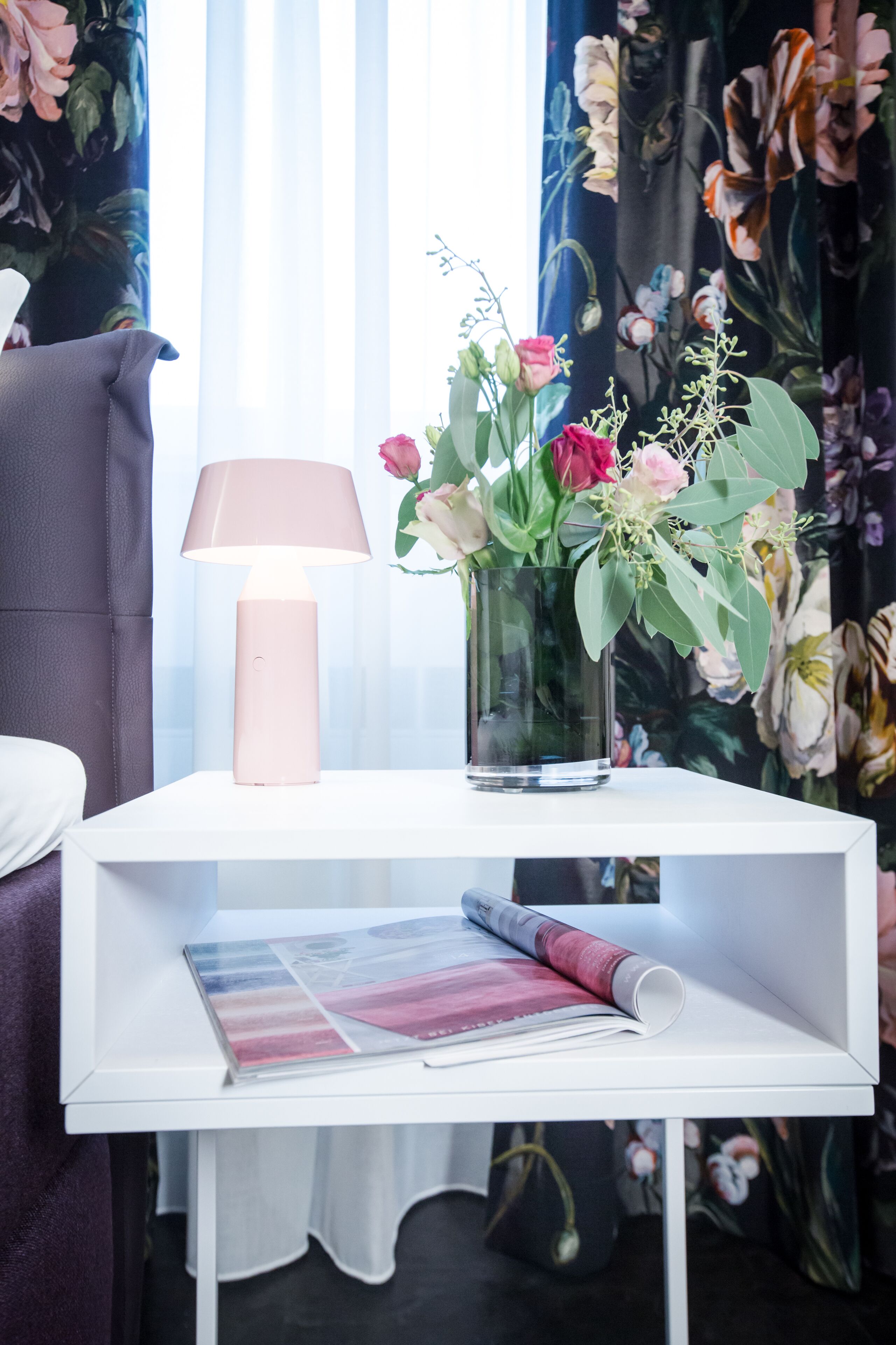 A white bedside table with a pink lamp, a vase with flowers, and a magazine.