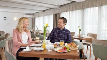 A couple dining in a modern hotel dining area with a large window, a vase with flowers, and a breakfast spread on the table.