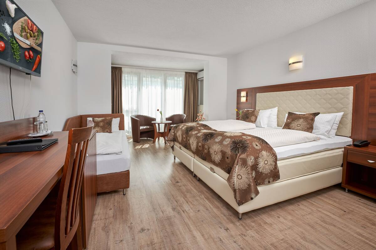 Quarto de hotel com uma cama grande, uma escrivaninha com cadeira, uma televisão e uma janela com cortinas.