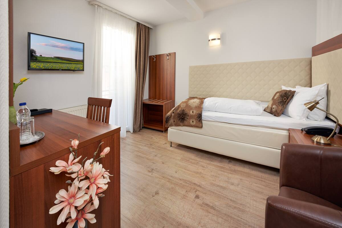 Quarto de hotel com cama, escrivaninha com cadeira, televisão e janela com cortinas.