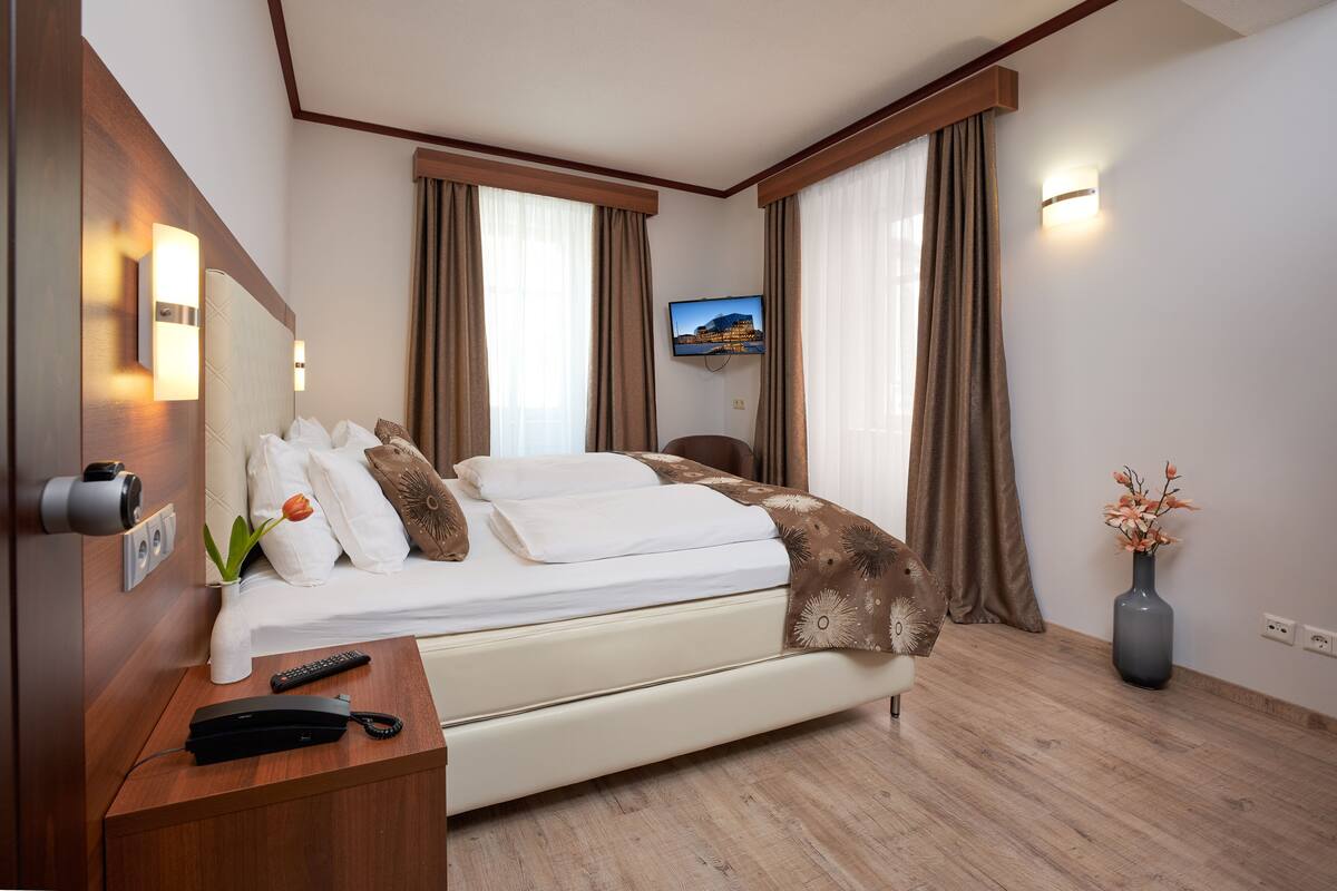 Quarto de hotel com uma cama, televisão presa na parede, escrivaninha com um controle remoto e um vaso com flores.