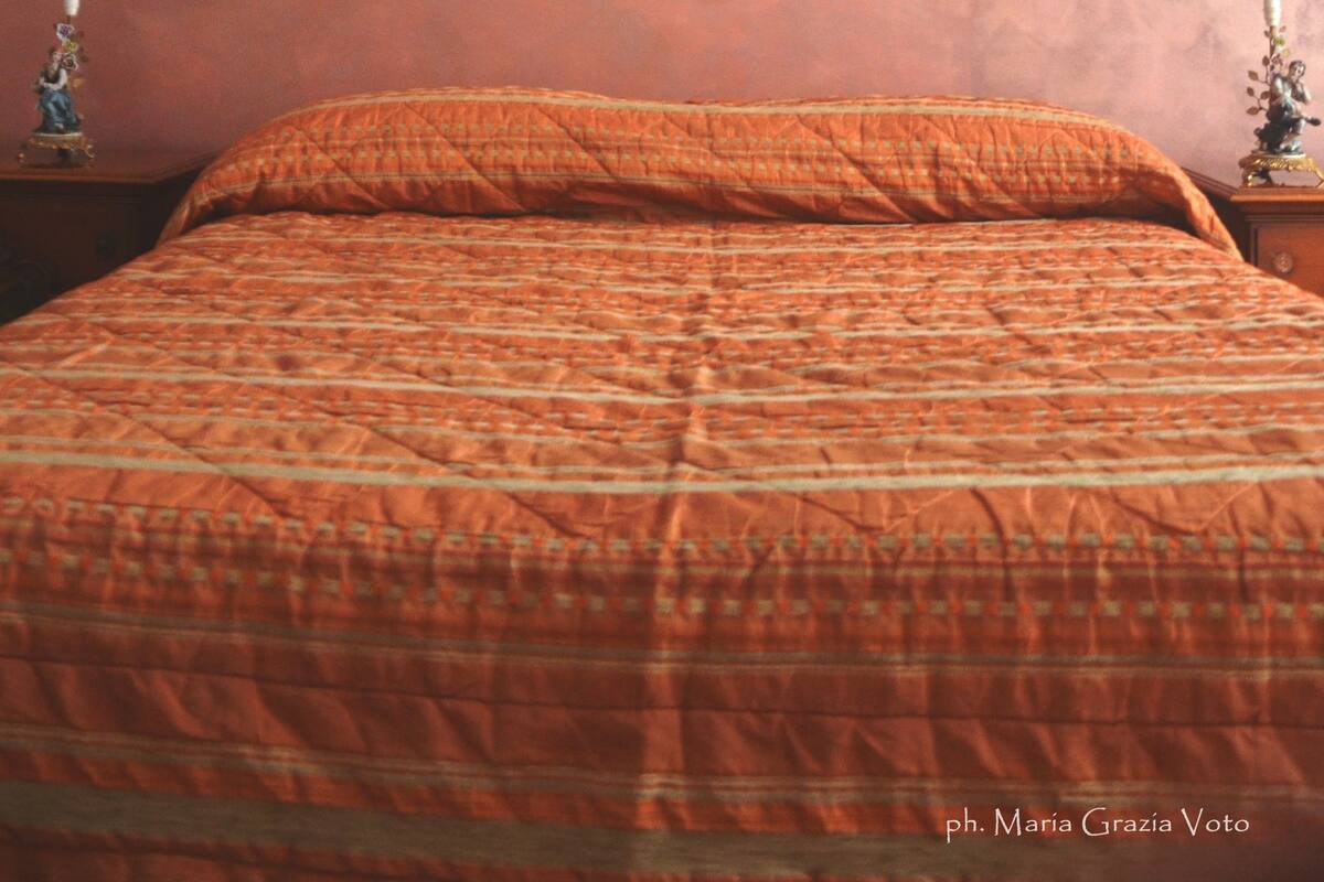 A bedroom with a bed, a wall-mounted artwork of a pink figure, and a patterned orange bedspread.