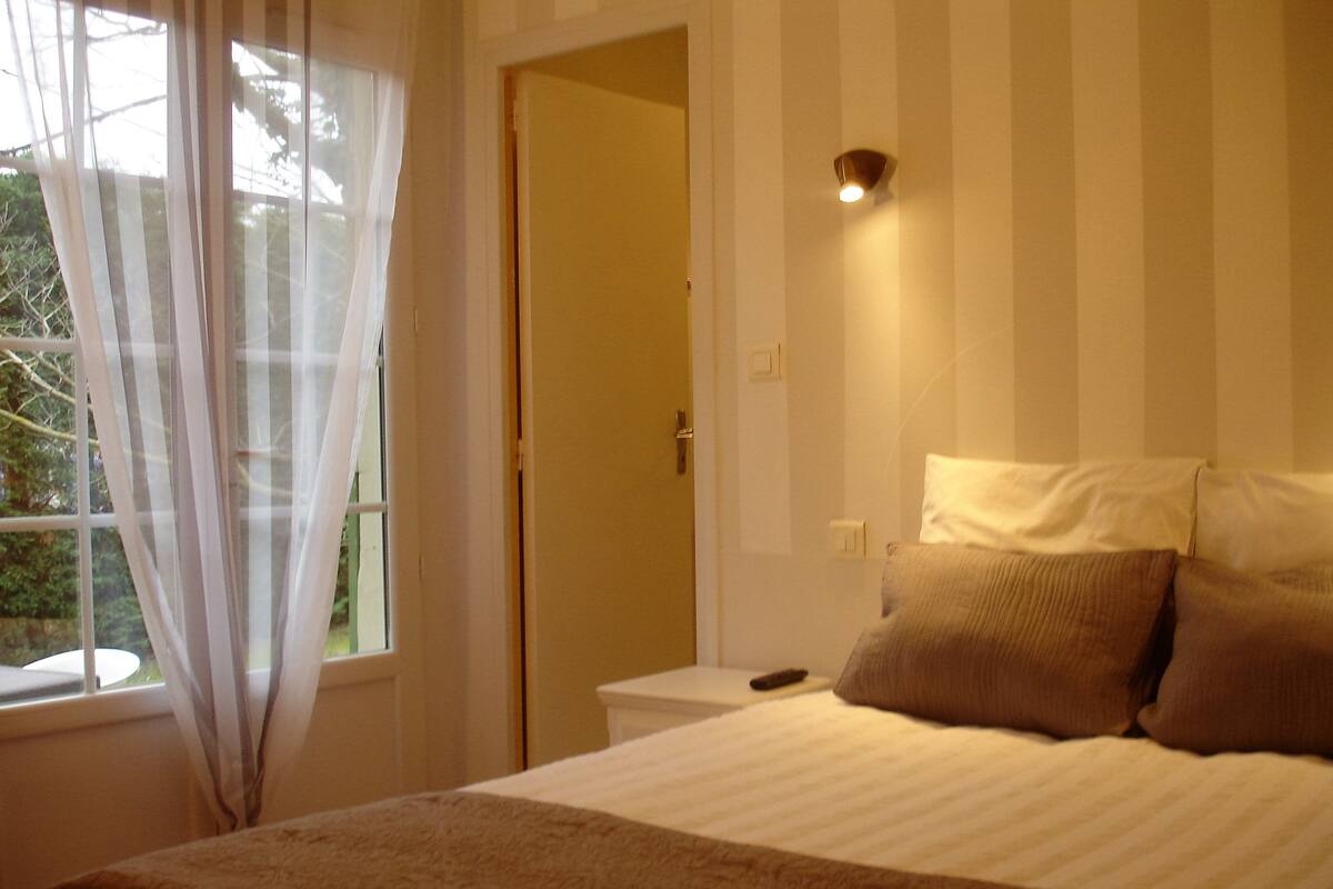 A bedroom with a bed, a bedside table, a nightstand, a window with curtains, and a door.