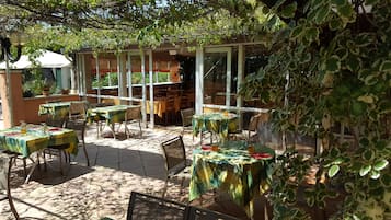 Un área de comedor al aire libre con mesas cubiertas con manteles a cuadros verdes y amarillos, rodeada de sillas y plantas en macetas.