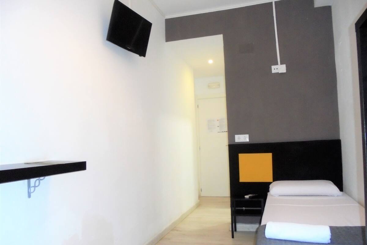 A hotel room with a bed, a bedside table, a flat-screen TV mounted on the wall, and a door leading to another room.