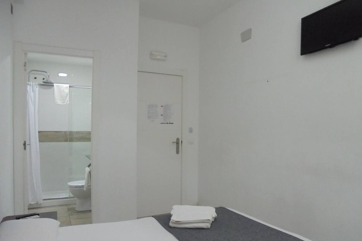 A hotel room with a bed, a bathroom with a shower, and a flat-screen TV mounted on the wall.