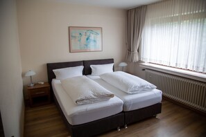 Ein Doppelbett mit weißen Bettwäsche, ein Nachttisch mit Lampe, ein gerahmtes abstraktes Gemälde an der Wand und ein Fenster mit durchsichtigen Vorhängen.