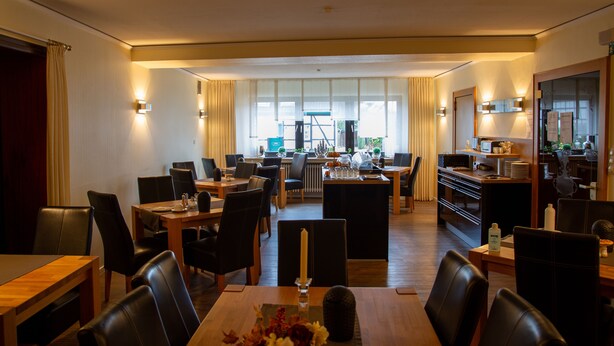 Une salle à manger d’hôtel avec de nombreuses tables et chaises, un îlot central doté d’un bar, et une vue sur la cuisine en arrière-plan.
