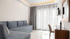 A modern living room with a grey sofa, a wooden side table, and a large window with sheer curtains. - 4-You (Polygyros)