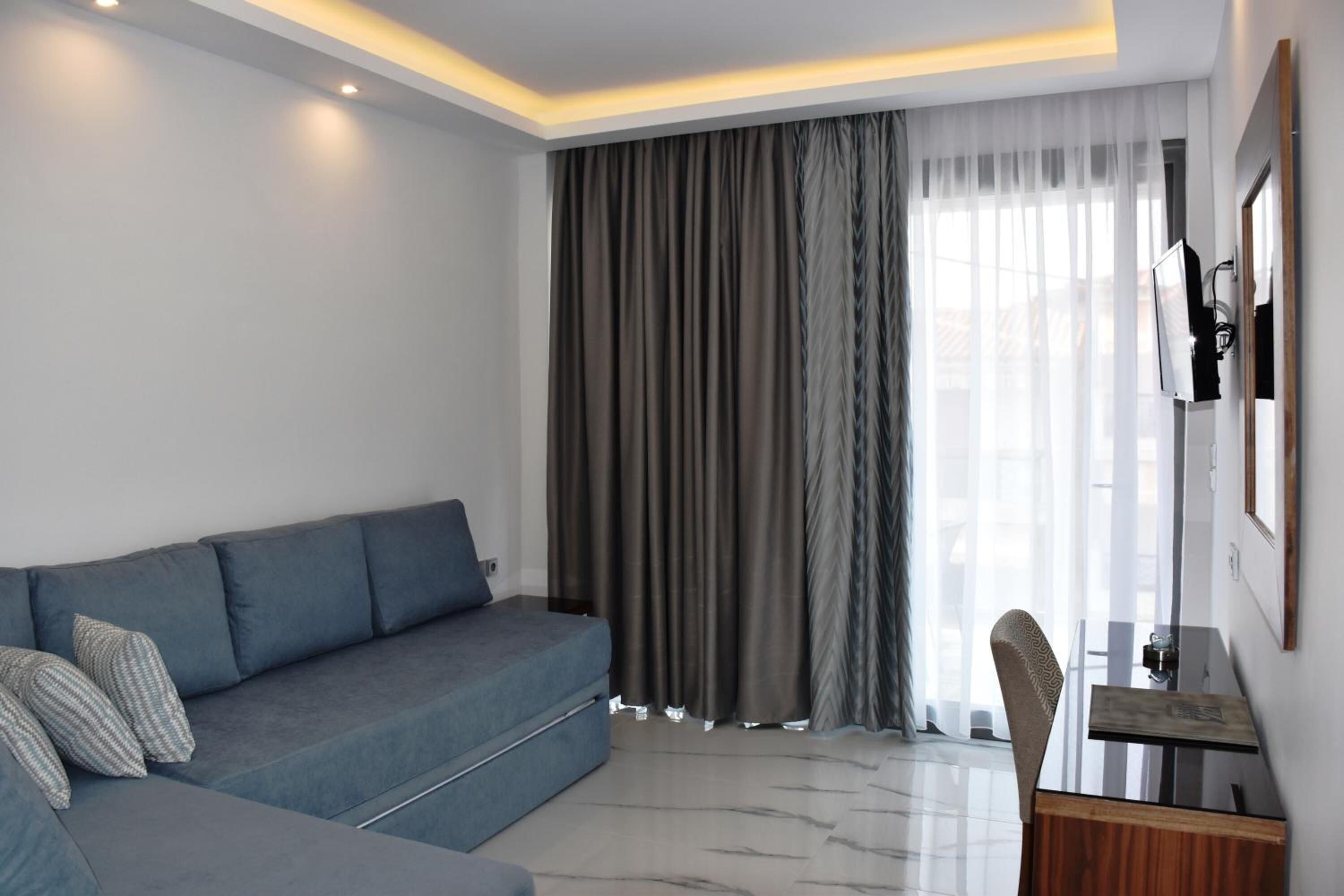 A modern living room with a grey sofa, a wooden side table, and a large window with sheer curtains.