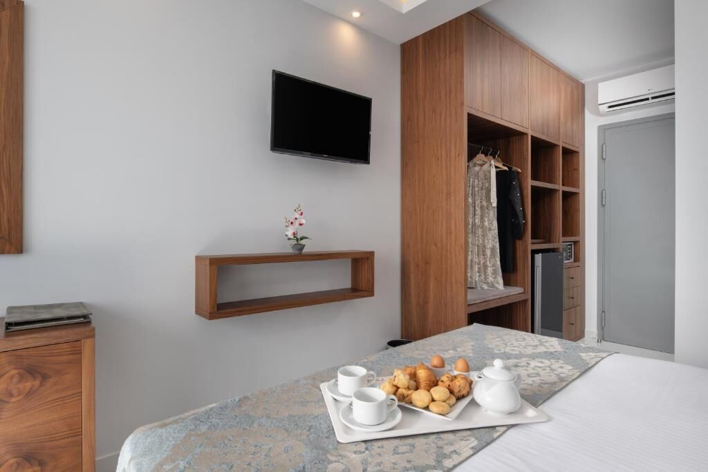 A hotel room with a bed, a TV mounted on the wall, a wooden wardrobe, a shelf with a potted plant, and a tray with breakfast items.