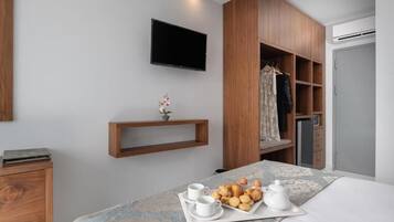 A hotel room with a bed, a TV mounted on the wall, a wooden wardrobe, a shelf with a potted plant, and a tray with breakfast items.