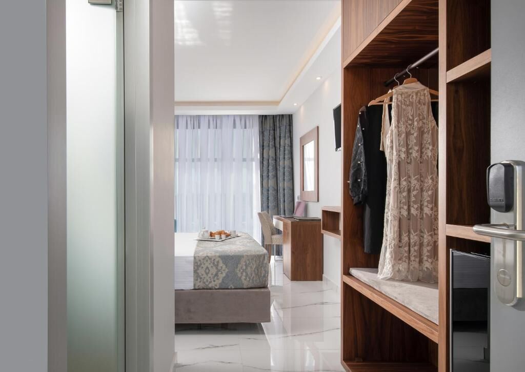 A modern hotel room with a wooden wardrobe, a bed with a patterned headboard, a small desk, and a view of the city through large windows.