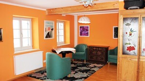 A room with orange walls, wooden flooring, teal chairs, a small table with a white tablecloth, and a cabinet with floral artwork. - Deutsches Haus (Waal)