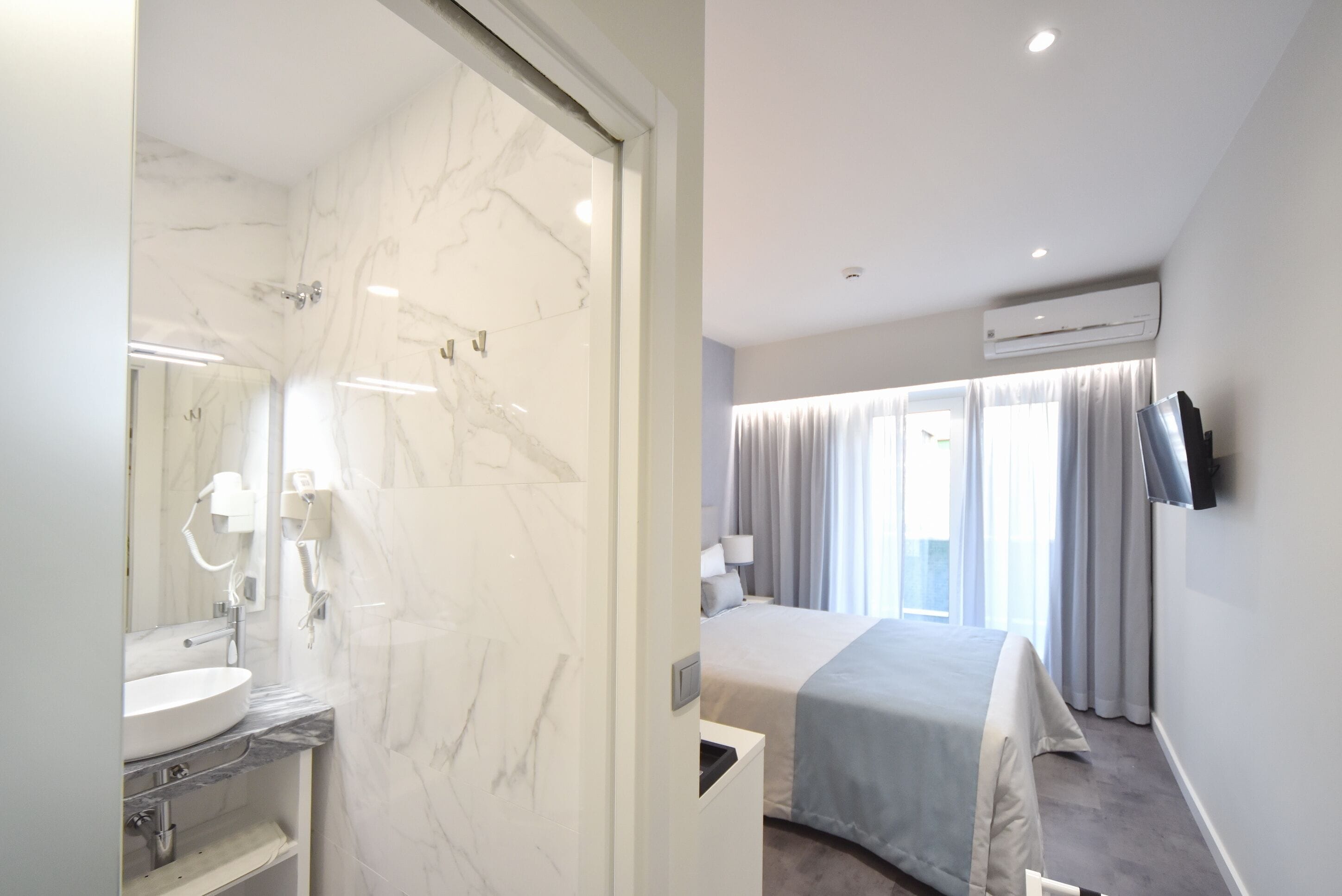 Quarto de hotel moderno com banheiro de mármore, cama com colcha cinza e televisão montada na parede.