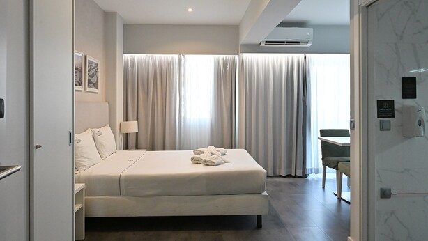 A modern hotel room with a bed, a desk, and a view of the outside through large curtains.
