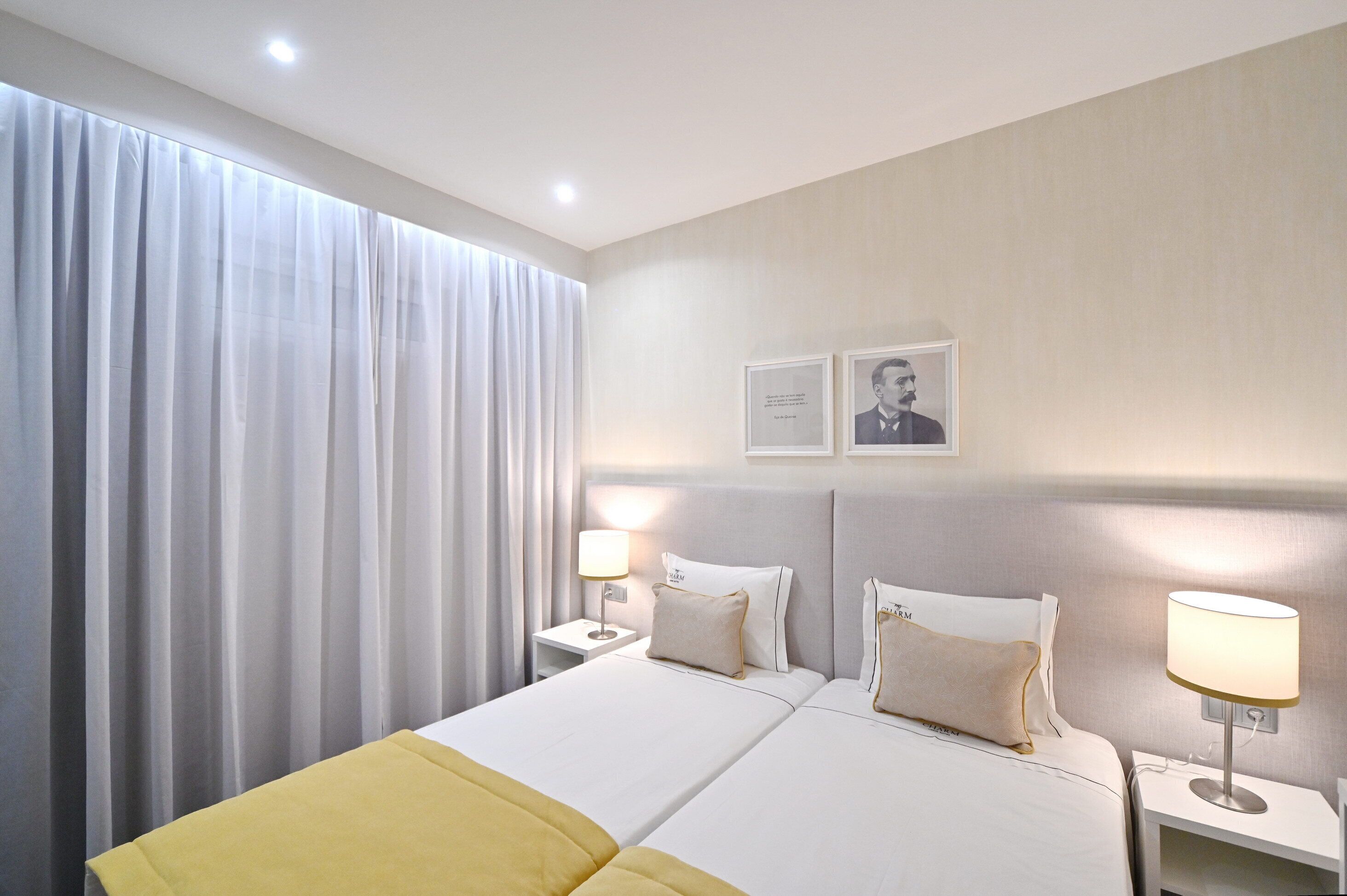A hotel room with two beds, a large window with curtains, bedside lamps, and framed pictures on the wall.