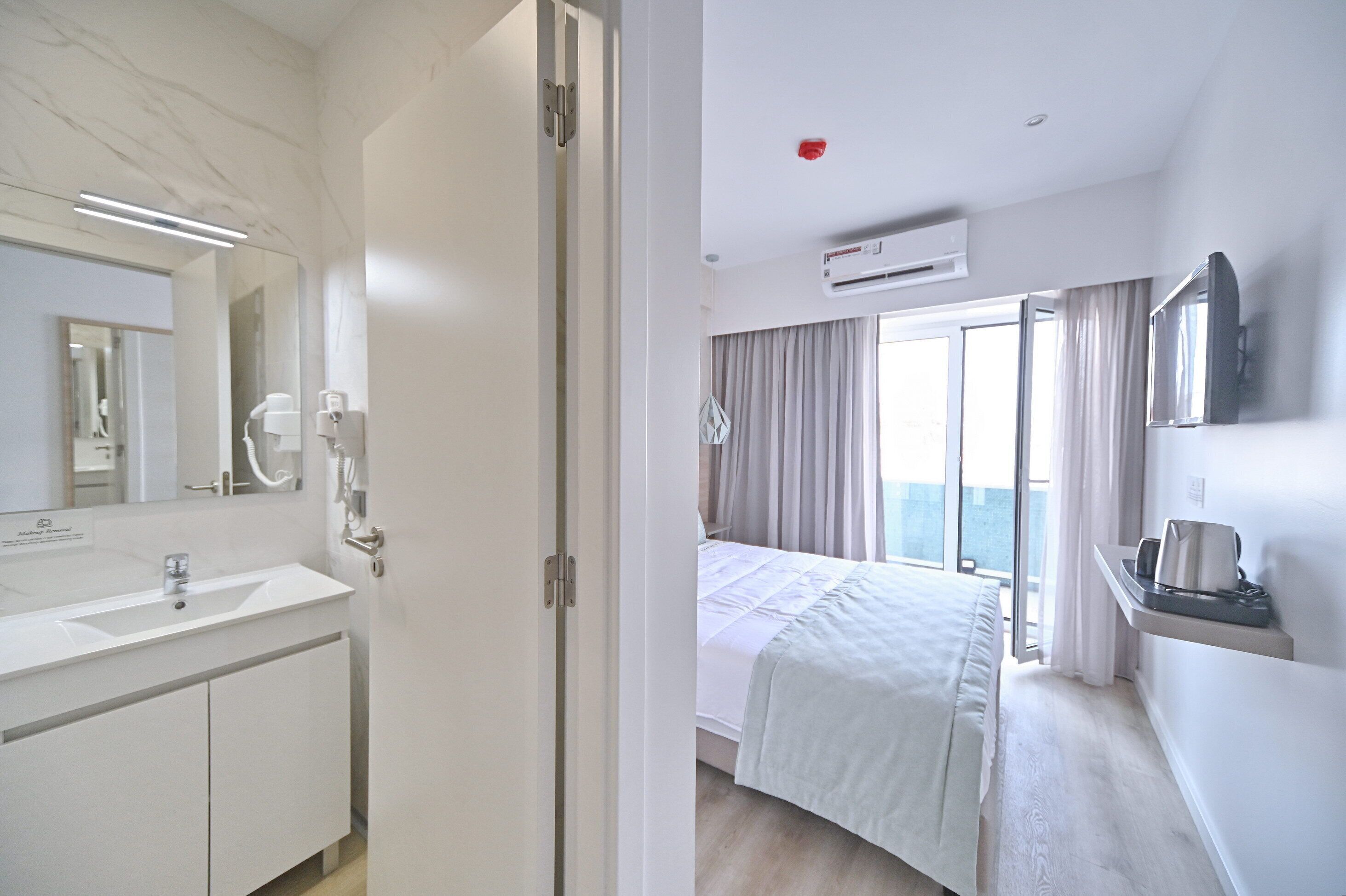 Habitación de hotel con un baño blanco, una cama con edredón blanco, una mesita de noche con hervidor y un ventanal con cortinas.