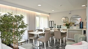 A modern dining area with white tables and chairs, a large potted plant, and a kitchen area with a refrigerator and microwave.