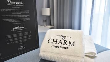 A hotel room with a neatly folded towel labeled "my CHARM LISBON SUITES" and a welcome card detailing the amenities and location of the My Charm - Lisbon Suites.