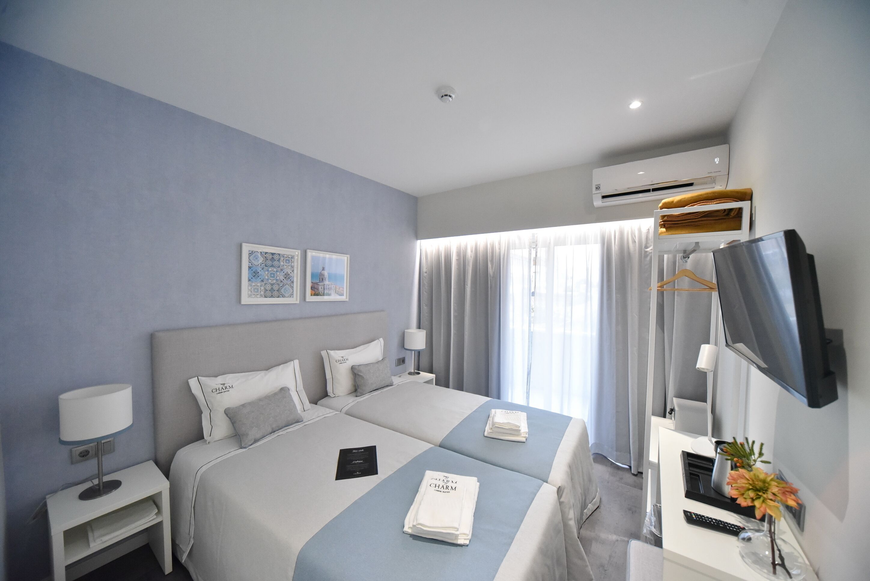 A hotel room with two beds, a flat-screen TV, a desk with a lamp, and a wall-mounted air conditioner.