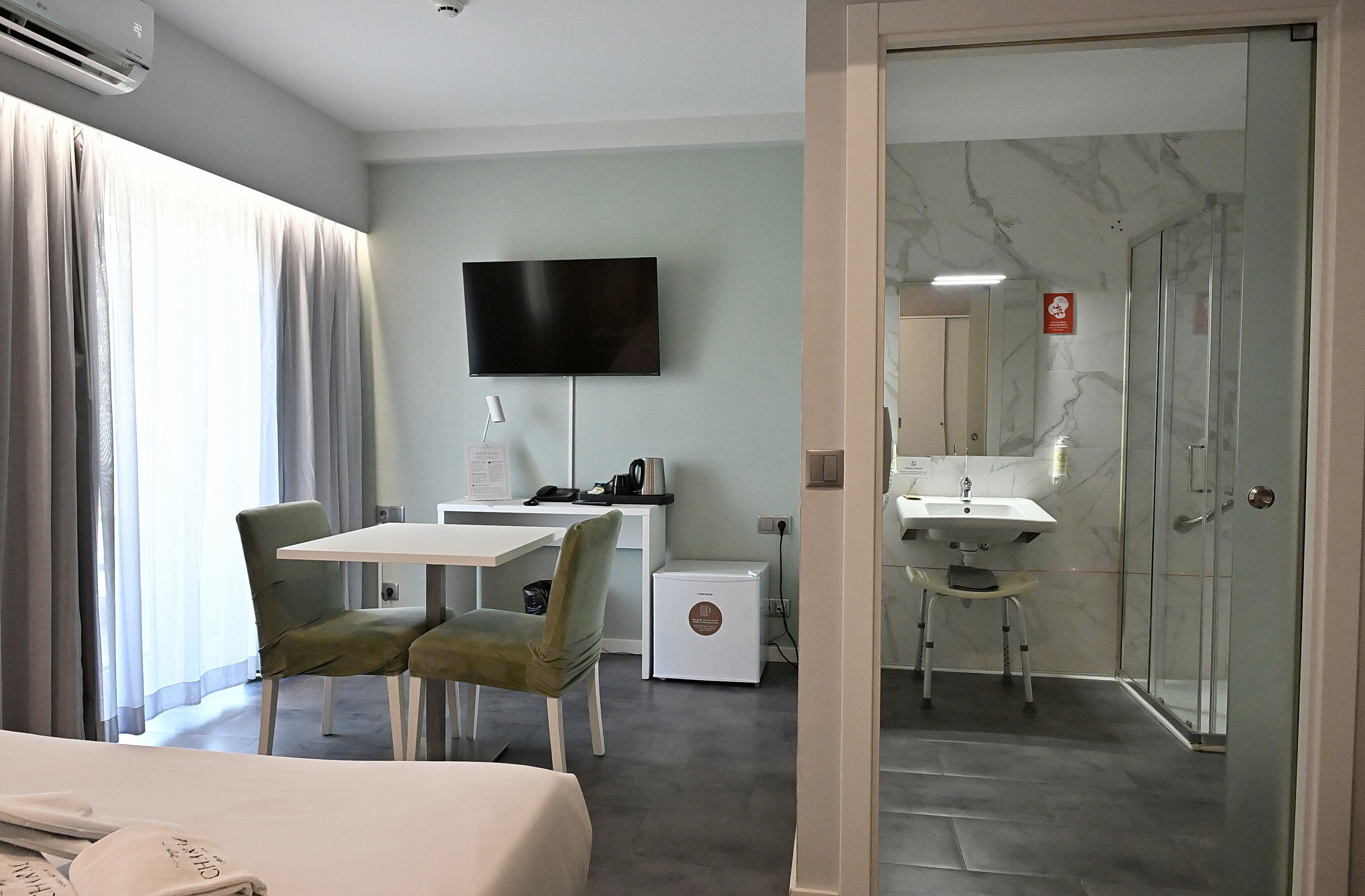 Quarto de hotel com cama, escrivaninha, cadeira, televisão, mini-geladeira e banheiro com chuveiro de vidro.