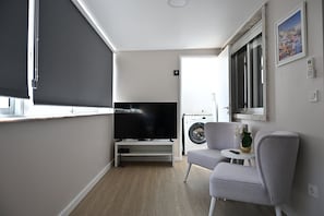 Ein modernes Wohnzimmer mit Flachbildfernseher, grauem Sofa, kleinem Tisch und einer Waschmaschine im Hintergrund.
