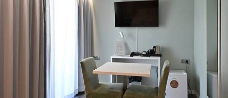 Una habitación con una televisión de pantalla plana en la pared, un escritorio blanco con una silla, un electrodoméstico pequeño y un amplio ventanal con cortinas.