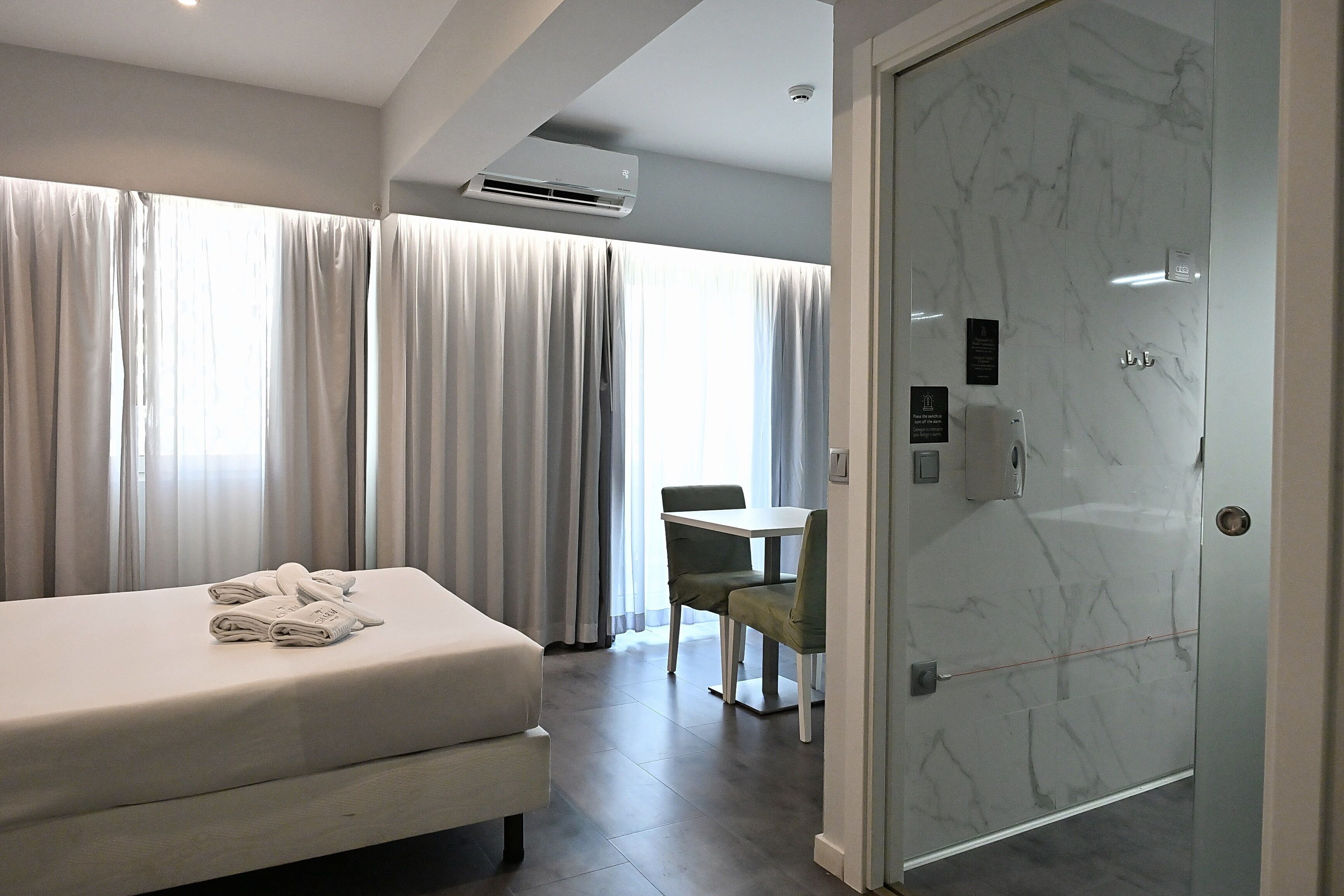 A hotel room with a bed, a table, and a chair. There is an air conditioning unit on the wall and a door with a handle.