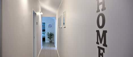 A hallway with a light-colored floor, white walls, and a door leading to another room. There are wall-mounted light fixtures and a potted plant.