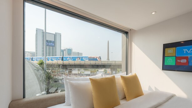 Ein Hotelzimmer mit großem Fenster, einem Bett mit weißen und gelben Kissen und einem an der Wand montierten Fernseher.