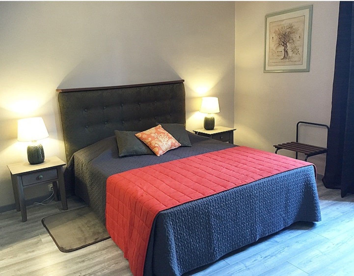 Une chambre à coucher avec une tête de lit sombre, une couvre-lit rouge matelassée, deux lampes de chevet et un tableau encadré au mur.