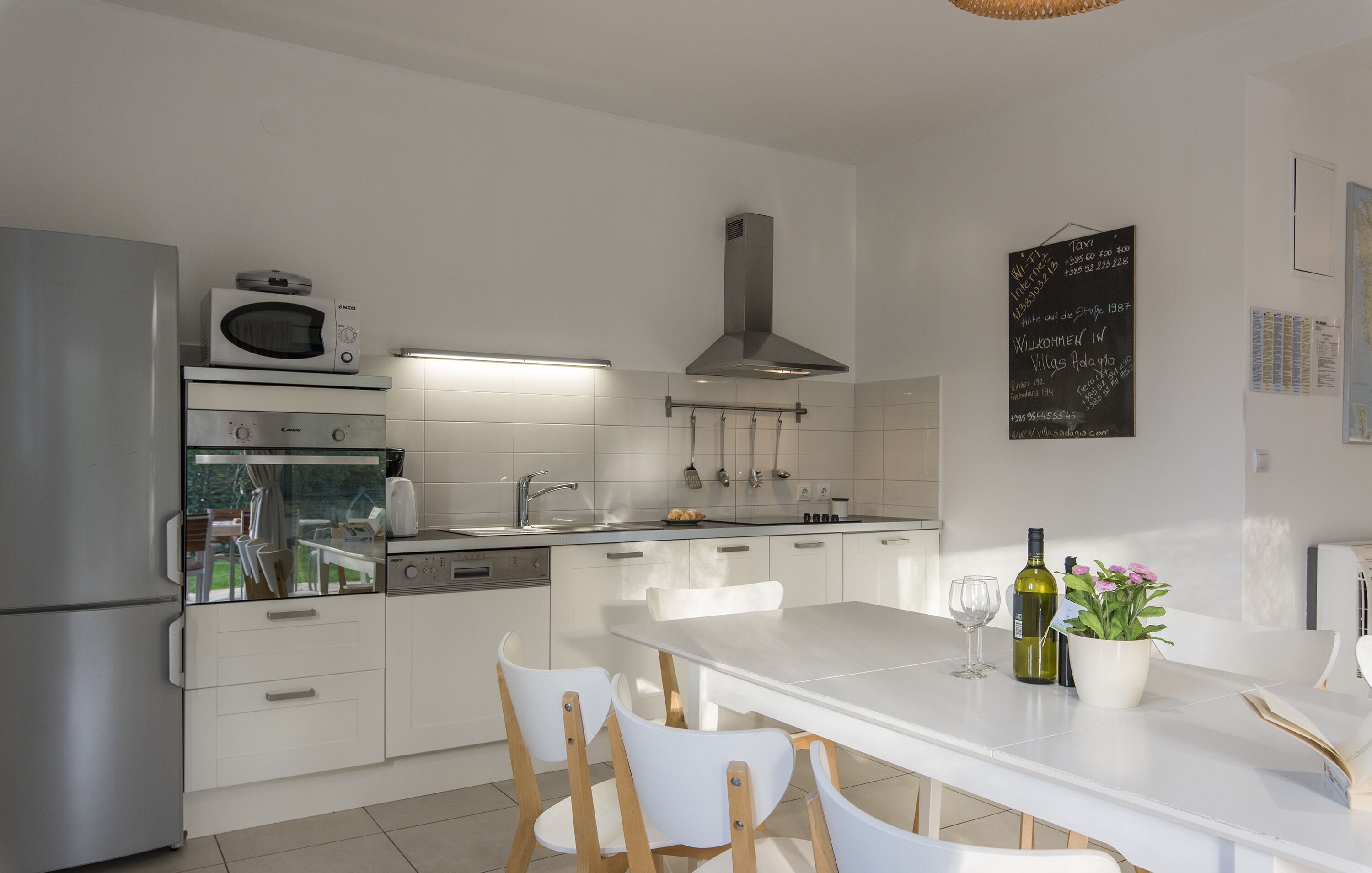 Eine moderne Küche mit weißen Schränken, einem Edelstahl-Kühlschrank, einer Mikrowelle und einem eingebauten Backofen. Ein Esstisch mit Stühlen, eine Weinflasche und ein Blumentopf sind ebenfalls vorhanden.