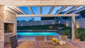 An outdoor patio with a wooden table and chairs, a stone fireplace, and a swimming pool. - Villas Adagio (Barban)