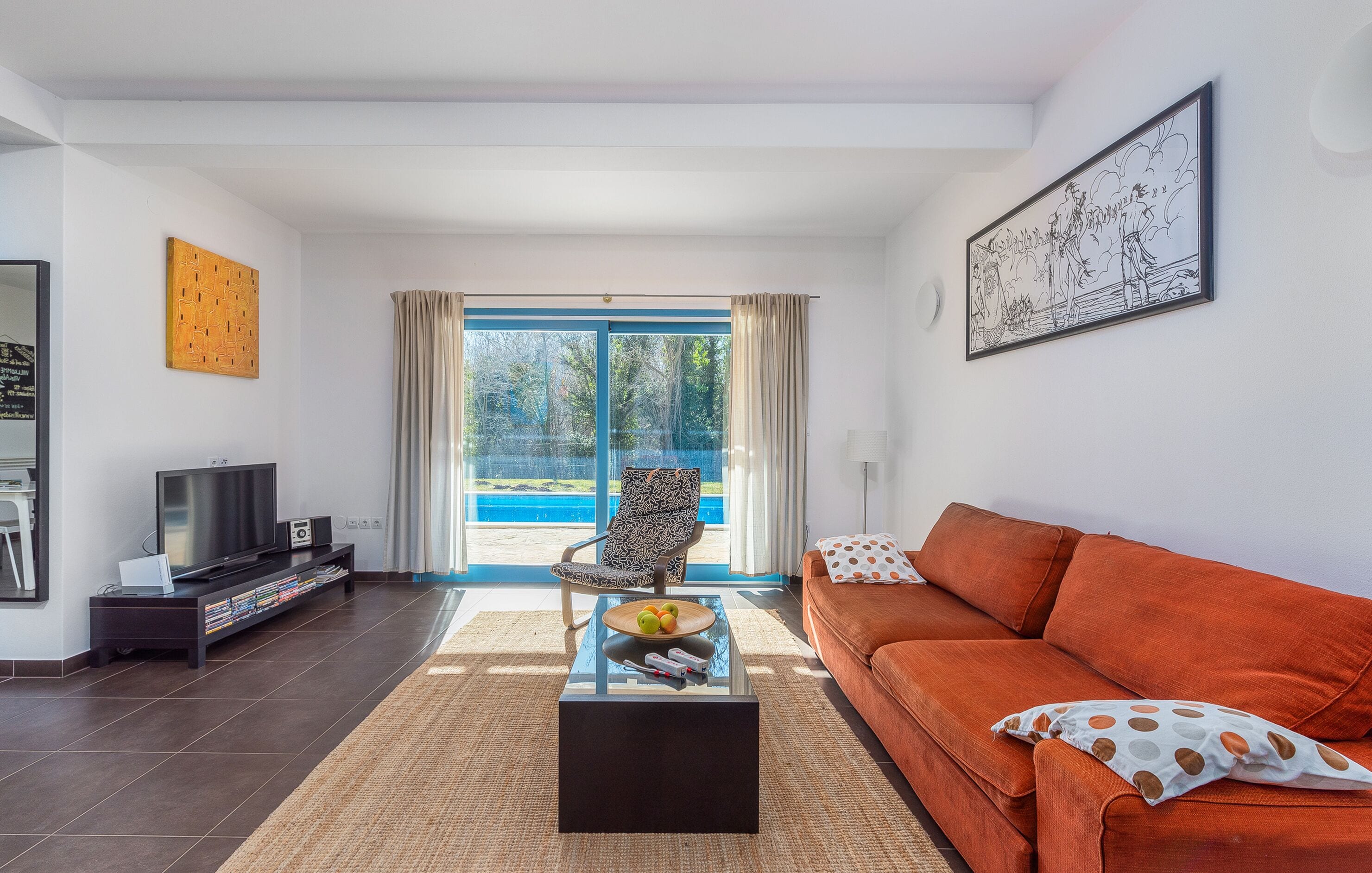 En moderne stue med sofa, sofabord, fjernsyn og en lænestol ved et stort vindue med udsigt til en pool.