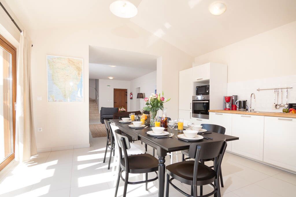 A modern kitchen with white cabinets, a dining area with a long table, and a view of the living room through an open doorway.