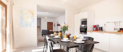 A modern kitchen with white cabinets, a dining area with a long table, and a view of the living room through an open doorway.