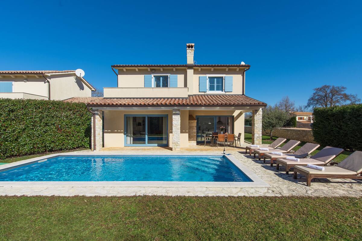 A two-story house with a swimming pool, a patio with lounge chairs, and a garden.