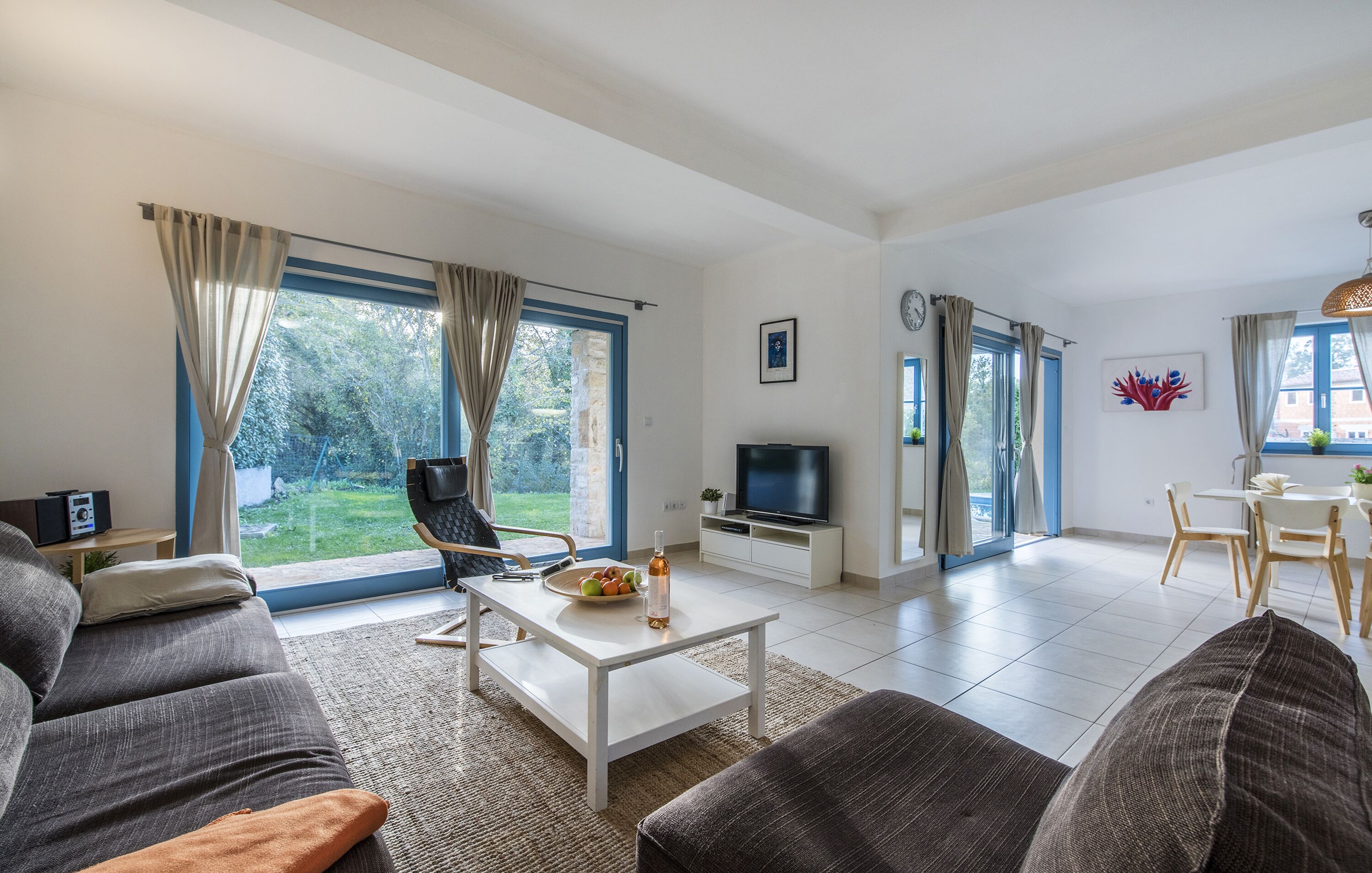 Een moderne woonkamer met een bank, salontafel en televisie. Er is een eethoek met een tafel en stoelen. Grote ramen bieden uitzicht op het buitengebied.