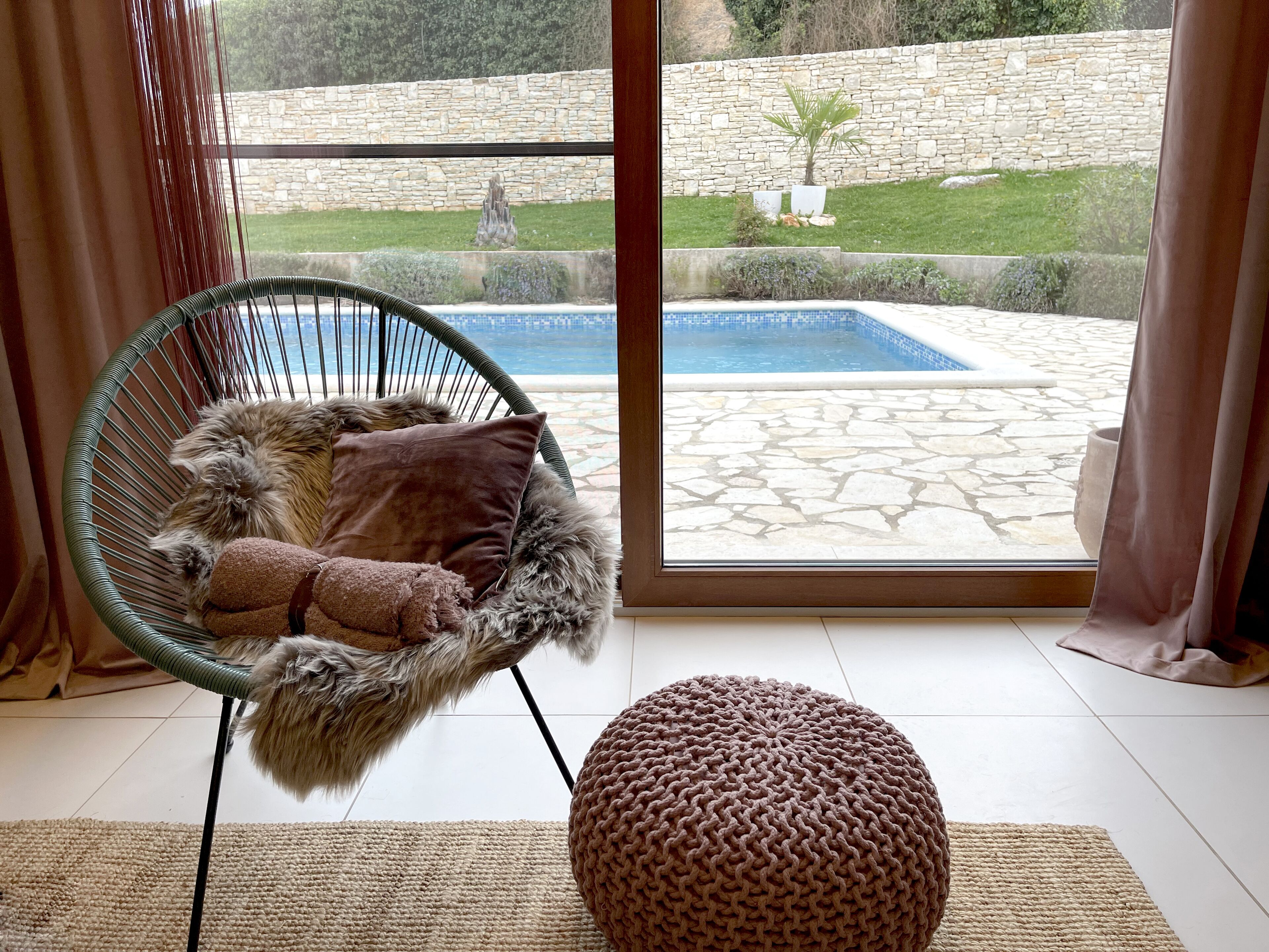 A room with a large window, a wicker chair with a fluffy cushion, and a knitted ottoman.