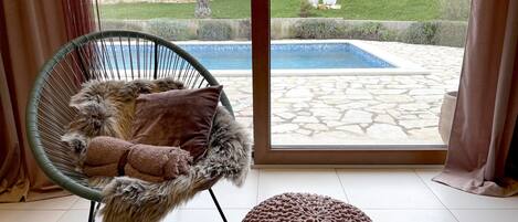 A room with a large window, a wicker chair with a fluffy cushion, and a knitted ottoman.