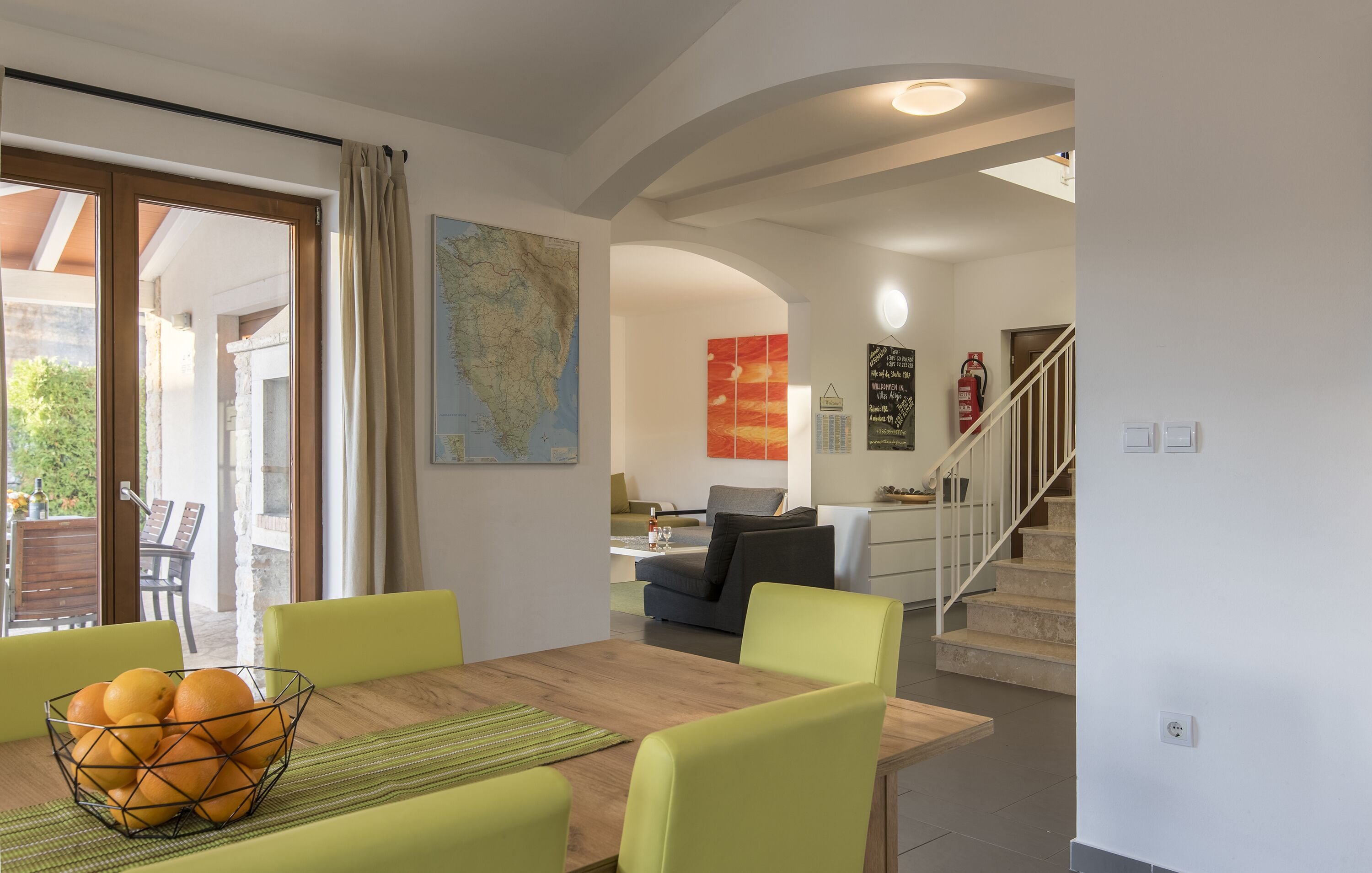 Un coin repas avec une table en bois, des chaises vertes et un saladier rempli d’oranges. Il y a une carte accrochée au mur et un escalier qui mène à une autre pièce.