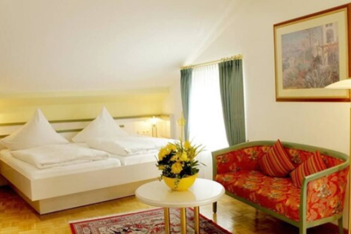 A hotel room with a bed, a sofa, a small table with a vase of flowers, a lamp, and a painting on the wall.