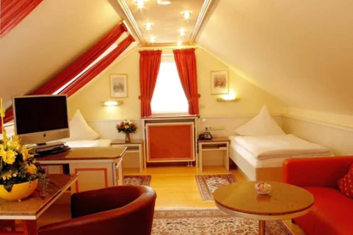 A cozy, well-lit attic room with a bed, a desk, a television, and a red sofa.